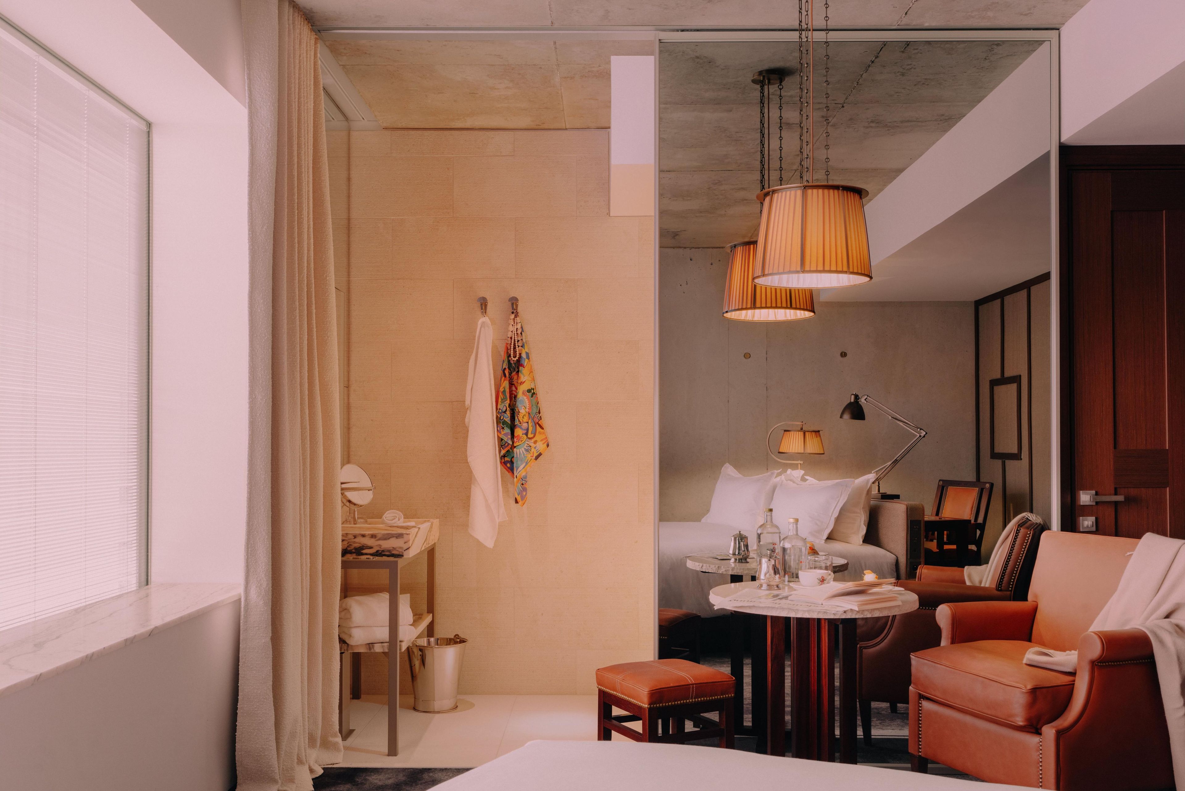 A hotel room with a bed, a round table with a lamp, a chair, and a small stool. There is a window with blinds, a curtain, and a wall-mounted lamp.