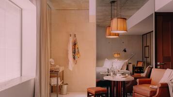 Habitación de hotel con una cama, una mesa redonda con una lámpara, una silla y un taburete pequeño. Hay una ventana con persianas, una cortina y una lámpara fijada a la pared.