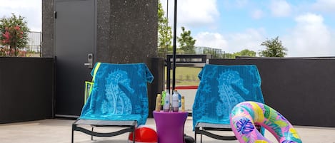 A poolside area with two lounge chairs, a blue umbrella, a pink lounge chair, a pool, and a colorful inflatable ring.