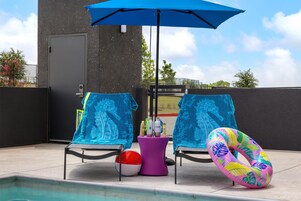 Zona de piscina con dos sillas de descanso, una sombrilla azul, una silla de descanso rosa, una piscina y un flotador inflable colorido.