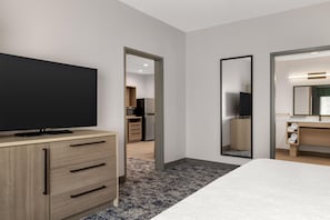 A hotel room with a bed, a TV on a wooden dresser, a bathroom with a mirror, and a kitchenette in the background.