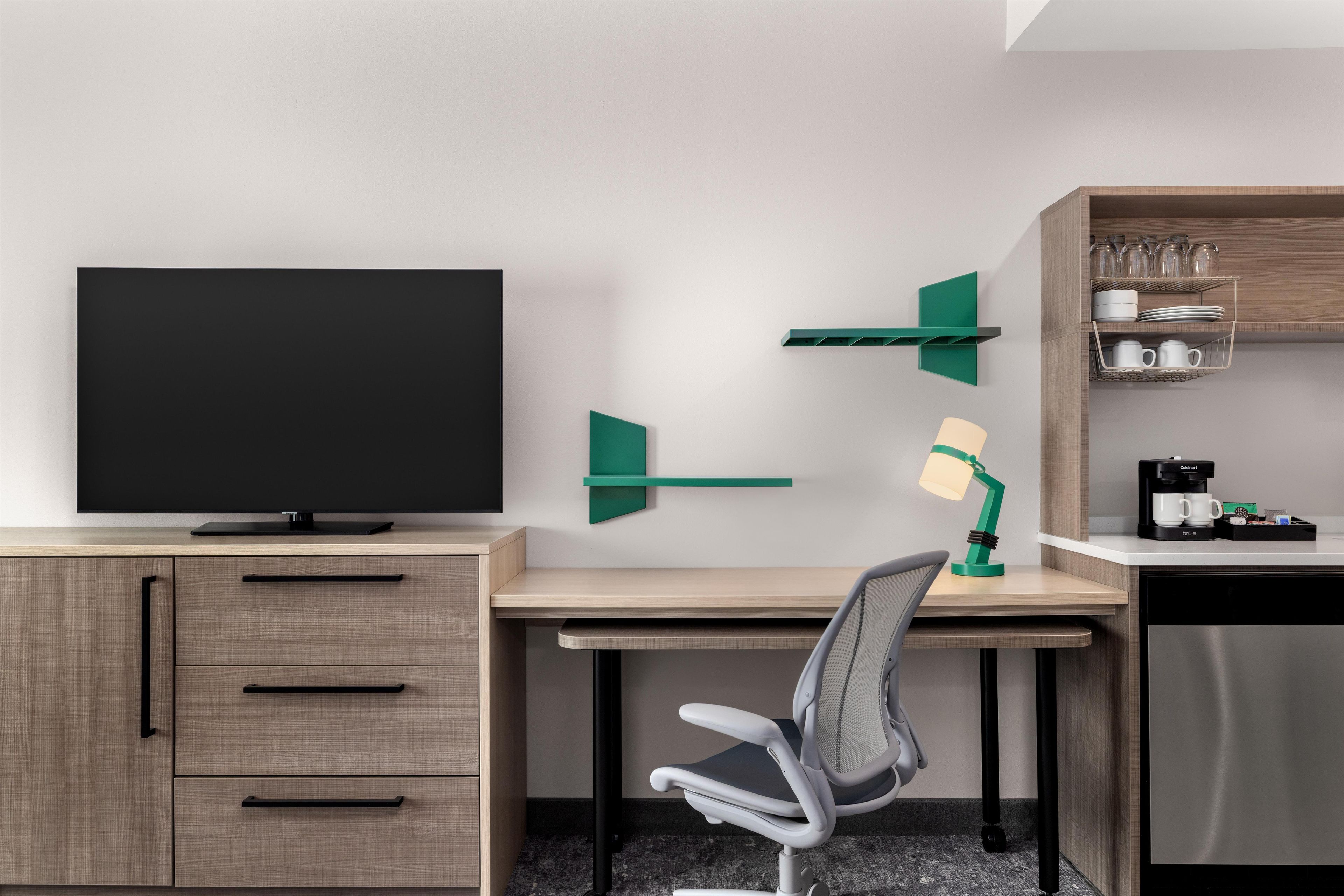 Habitación de hotel moderna con televisión de pantalla plana, un escritorio con silla, cafetera y estante con utensilios de cocina.