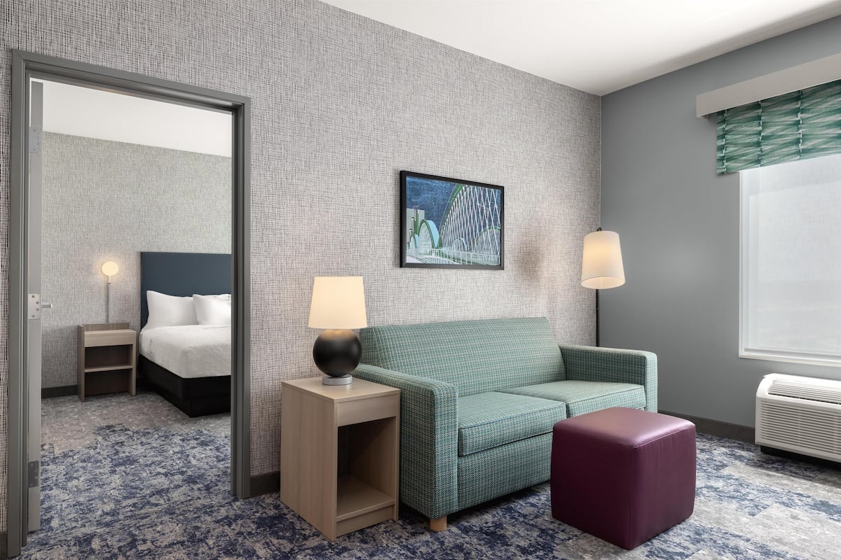 Une chambre d’hôtel avec un canapé vert, une petite table avec une lampe, un pouf violet, un lit avec du linge de maison blanc, un tableau encadré au mur et une fenêtre avec un rideau vert.