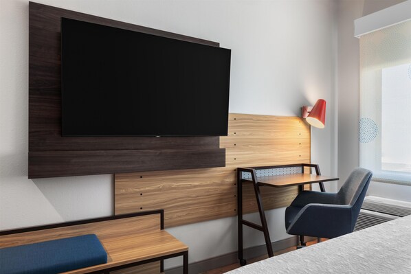 Ein modernes Hotelzimmer mit Flachbildfernseher an einer Holzwand, einem Schreibtisch mit Stuhl, einer Bank und einer roten Lampe.