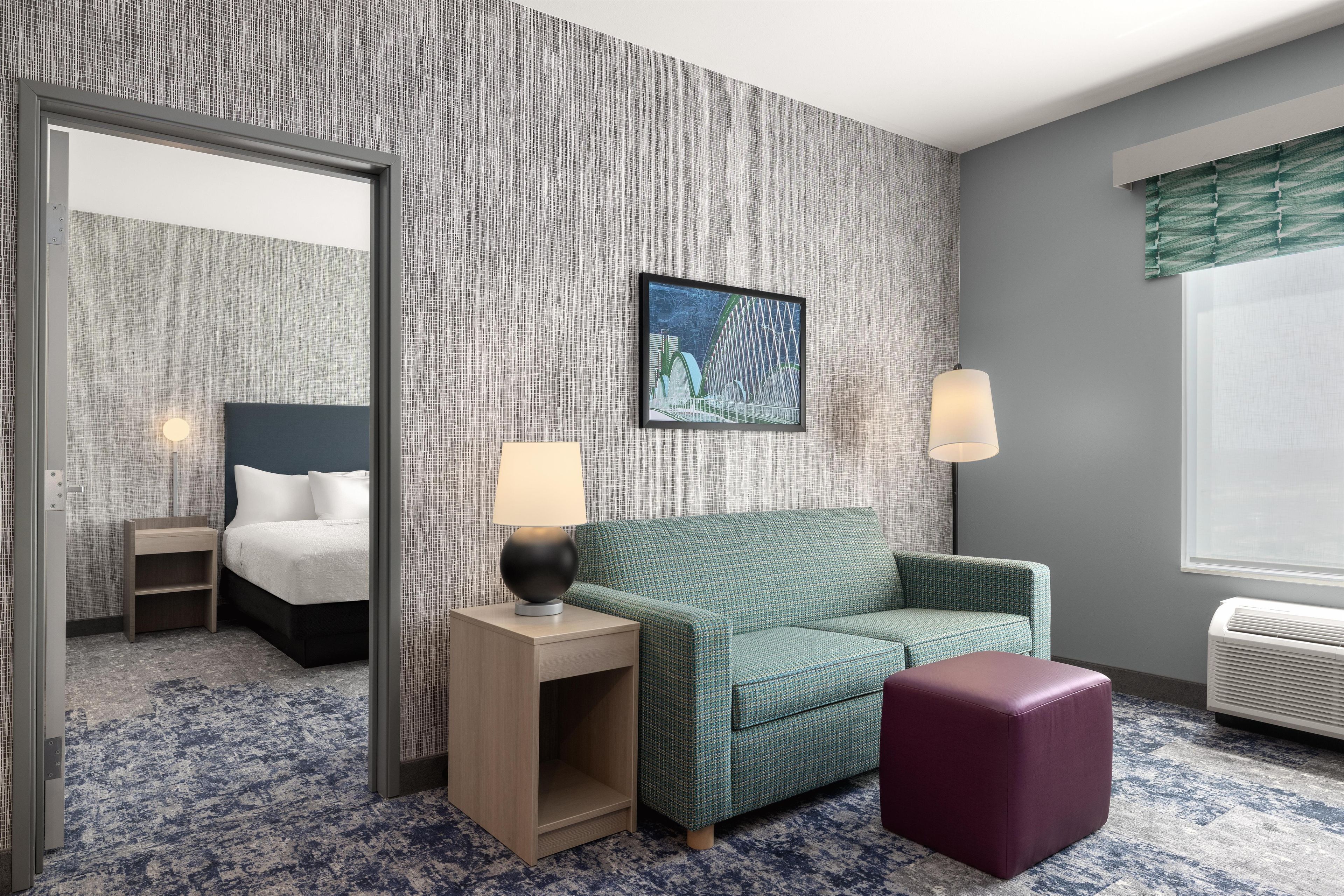 A hotel room with a bed, a green sofa, a small table with a lamp, a picture on the wall, and a window with a view.