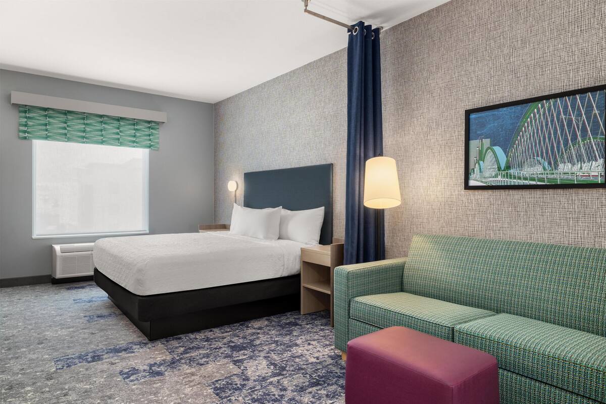 Une chambre d’hôtel avec un lit, un canapé vert, un pouf rouge, une table de chevet, une lampe et un tableau représentant un pont.