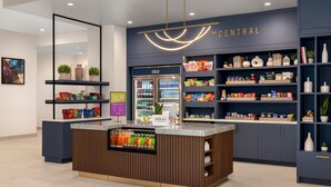 Eine moderne Hotelhalle mit einer zentralen Rezeption, einem Kühlregal für Getränke sowie Regalen voller Snacks und Bücher.