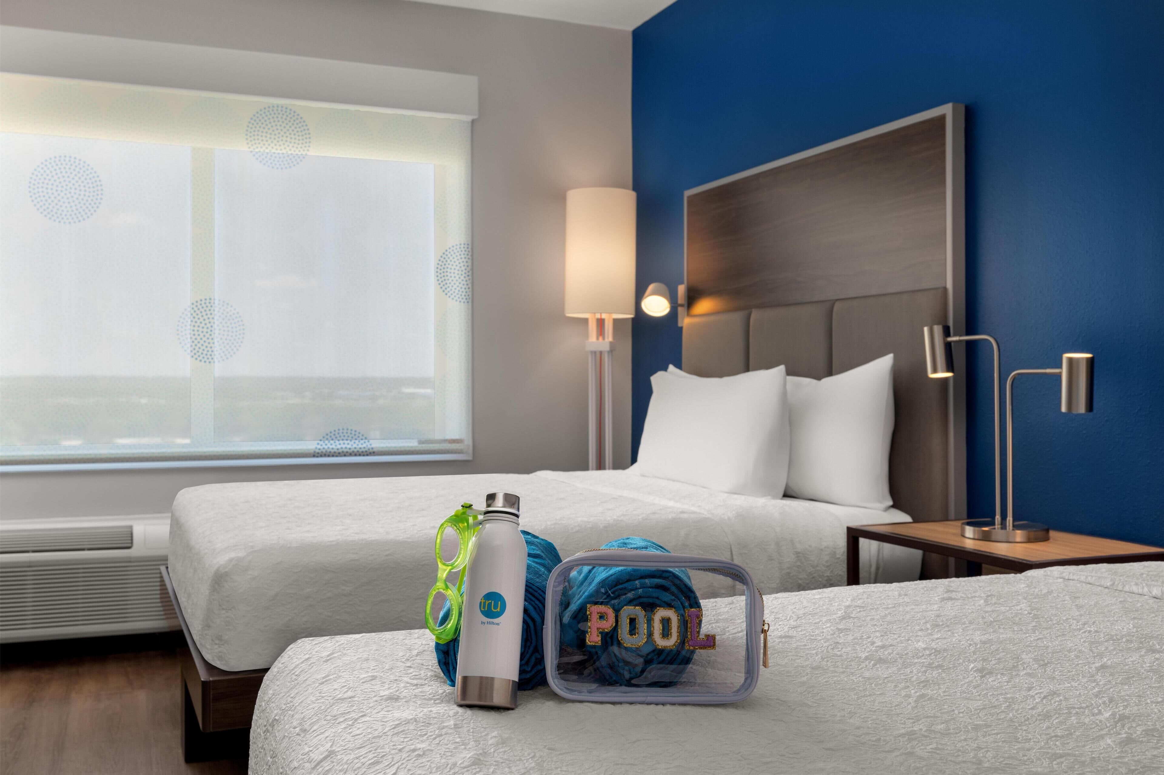 Una habitación de hotel con dos camas, una ventana con persianas, una lámpara de noche, una botella de agua, un par de gafas y una pelota azul con la palabra 'POOL' escrita.