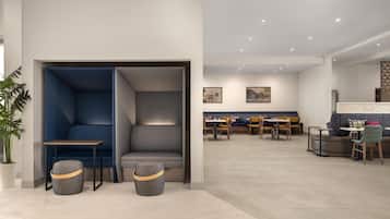 Een modern interieur met een zithoek, bestaande uit een blauwe bank, een klein tafeltje en twee poefs. Op de achtergrond staan meerdere tafels en stoelen, wat wijst op een eet- of vergaderruimte.