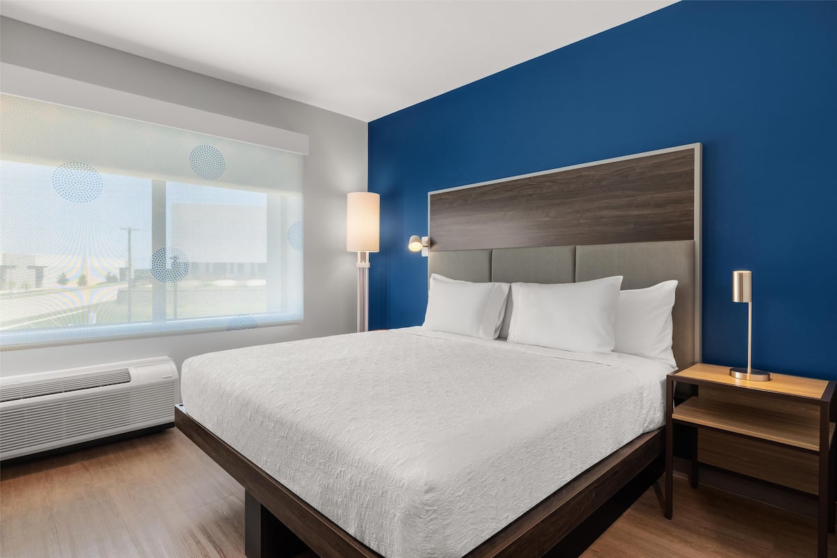 A modern hotel room with a large bed, a bedside table, a lamp, and a view of the city through a large window.