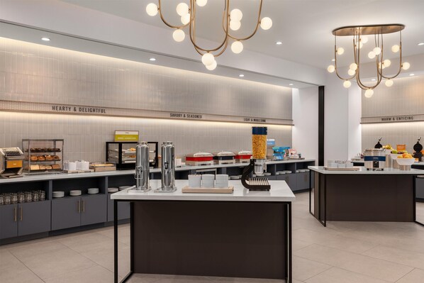 Un área de desayuno moderna con una isla central, distintas estaciones de comida y un diseño limpio y contemporáneo.