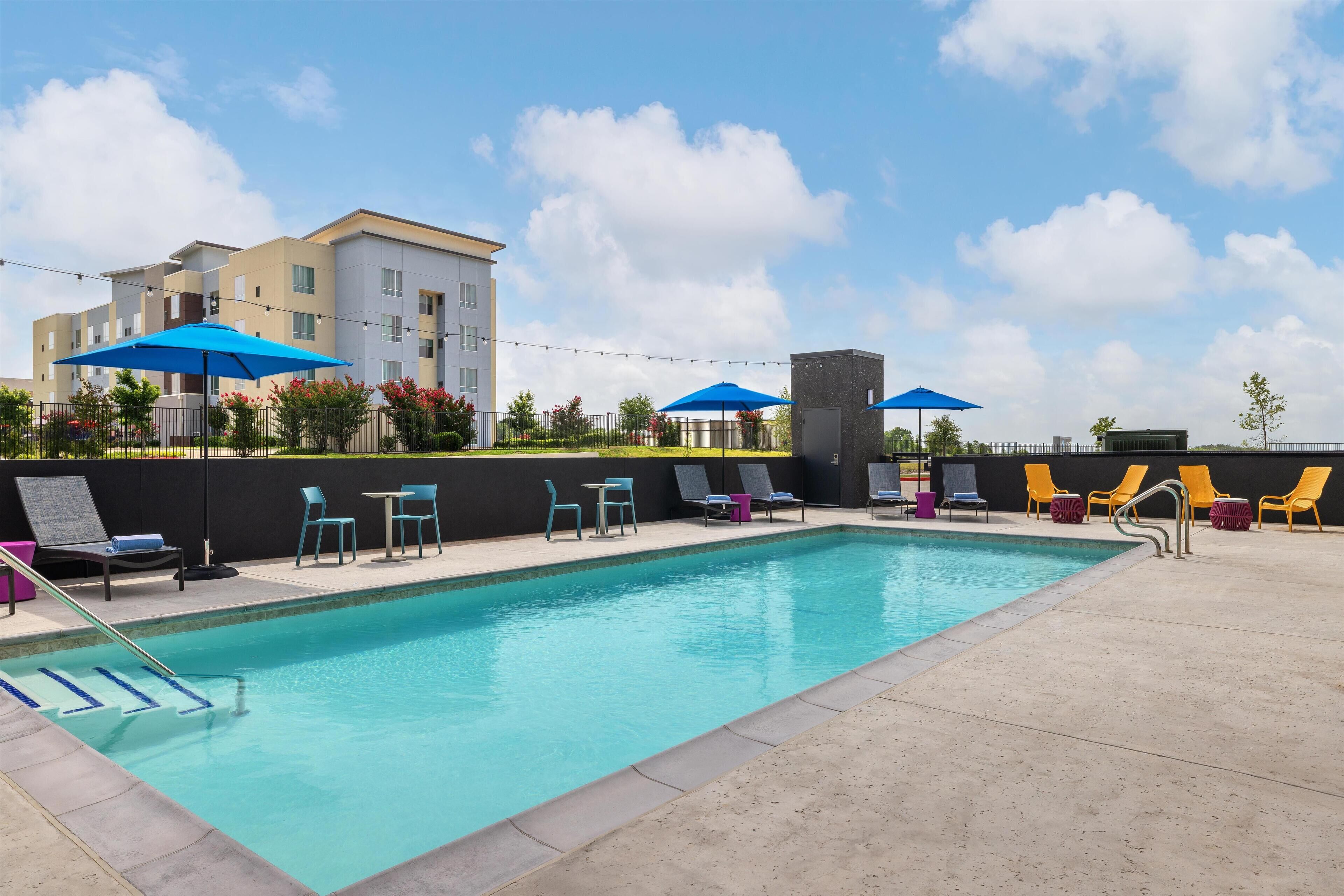 A swimming pool with lounge chairs and umbrellas, surrounded by a building with multiple floors.