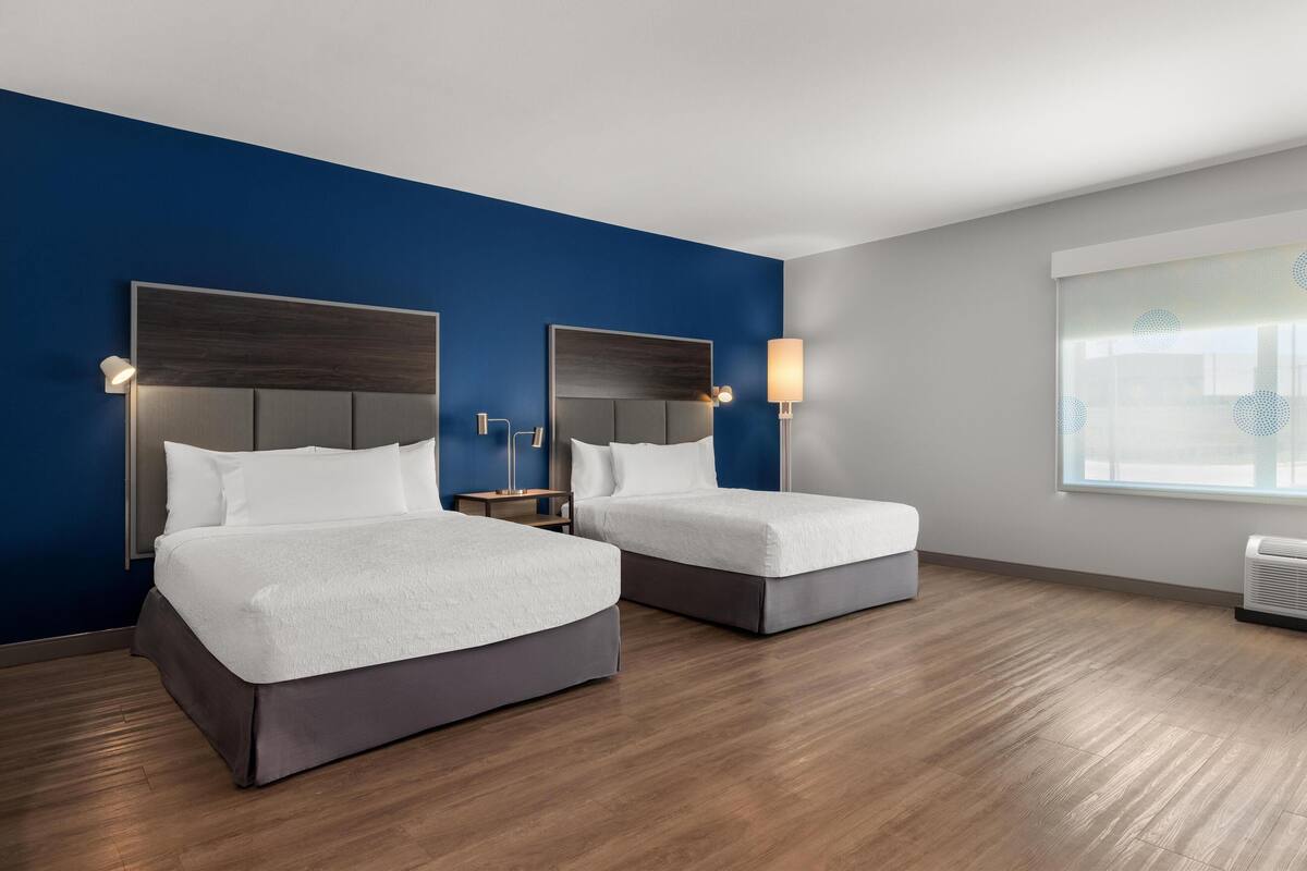 A hotel room with two beds, a blue accent wall, a window with blinds, and a wooden floor.