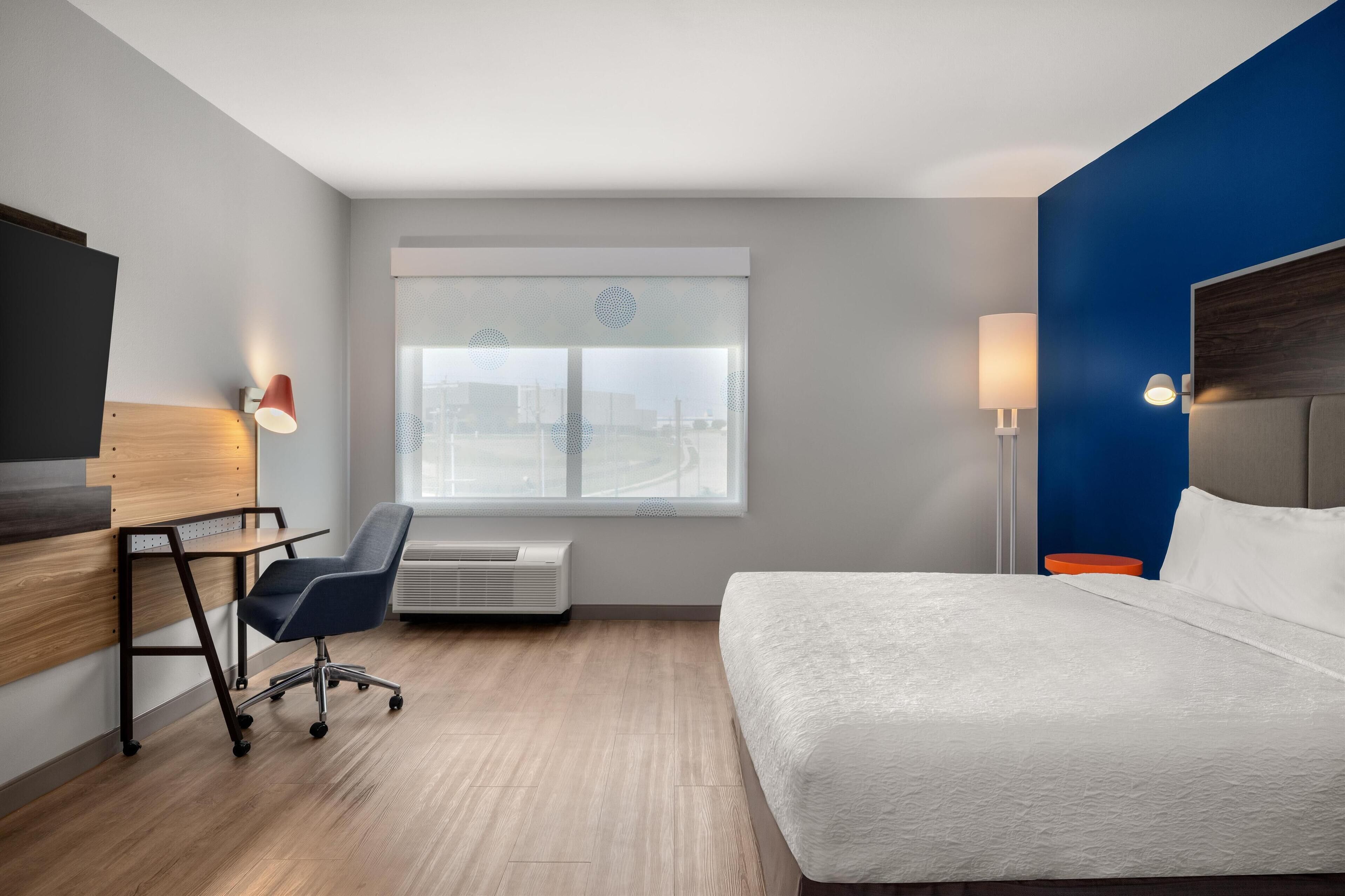 A hotel room with a bed, desk, chair, and a window with a view of a cityscape.