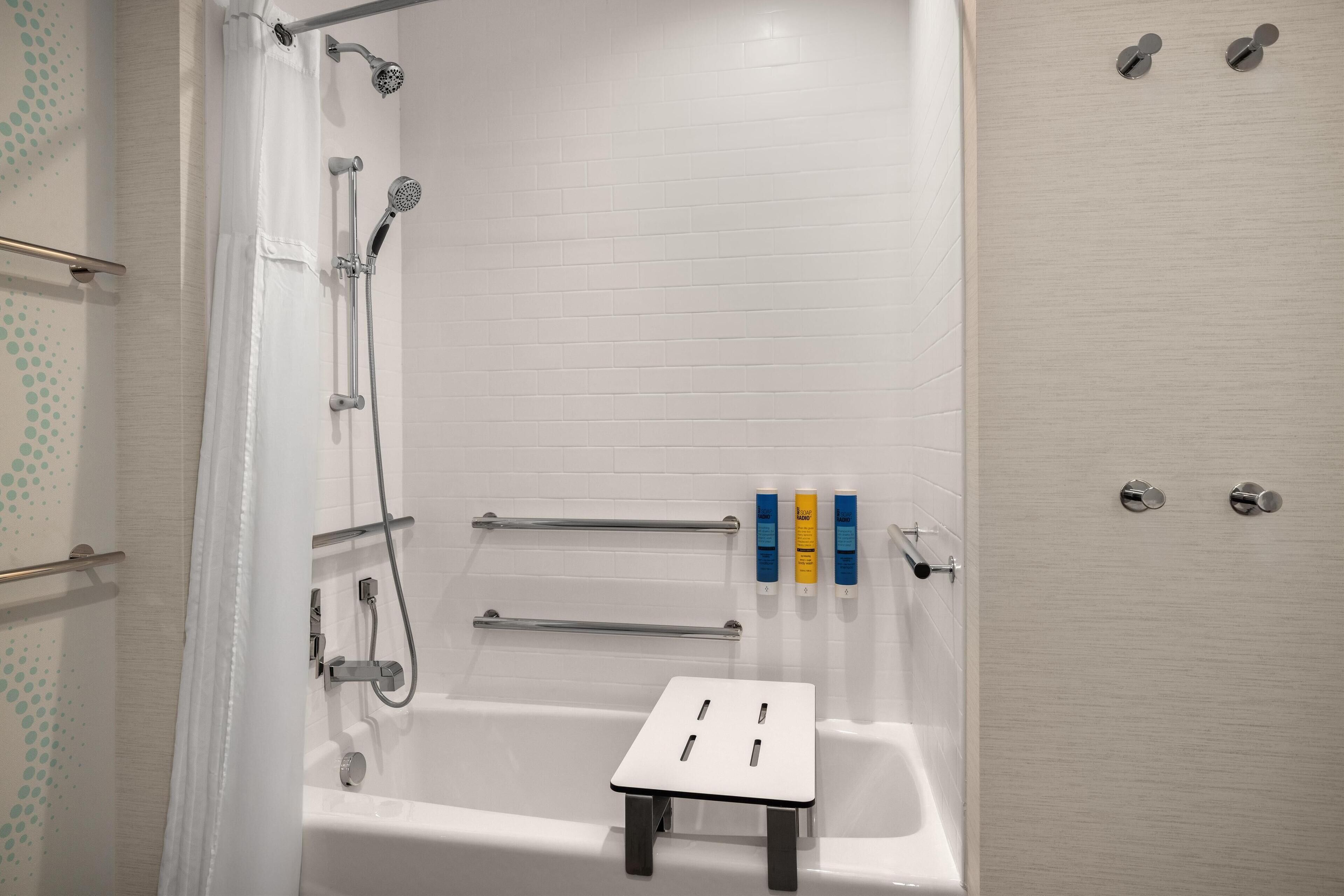 Un baño con bañera, ducha y barras de apoyo. Hay artículos de aseo en la pared.