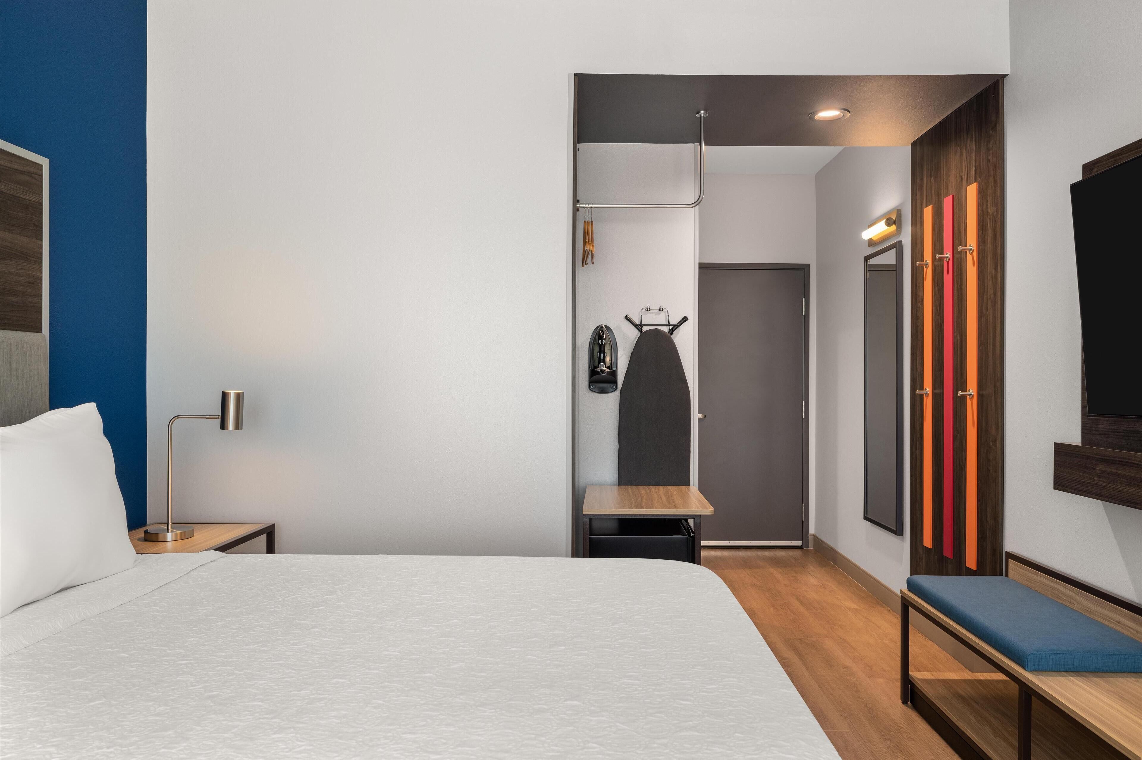 Habitación de hotel moderna con cama, mesita de noche, lámpara, banco y espejo con televisor.