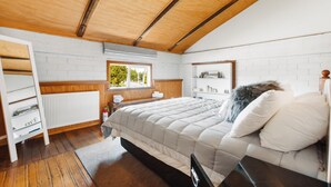 A bedroom with a wooden ceiling, white brick walls, a bed with a white duvet, a window with a view of greenery, and a white radiator. - The Workshops - Clyde Holiday Home (Clyde)