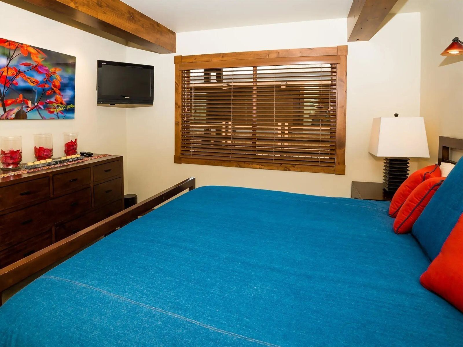 A bedroom with a large bed, wooden headboard, a TV mounted on the wall, a window with blinds, and a bedside lamp.