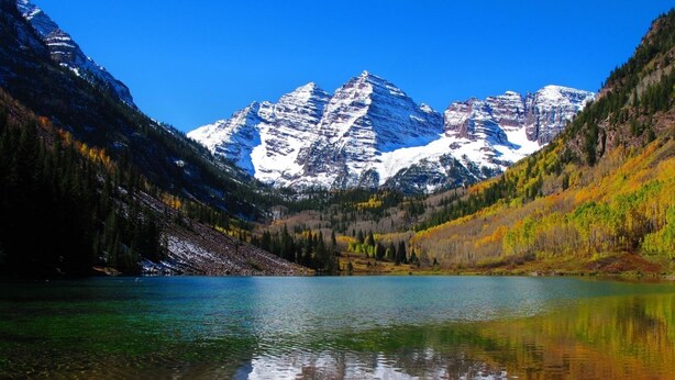 Un lago de montaña con reflejos cristalinos, rodeado de laderas boscosas y cumbres nevadas.