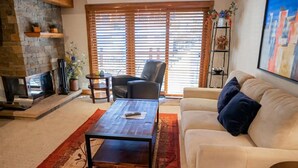 A living room with a fireplace, a sofa, a coffee table, and a large window with blinds.