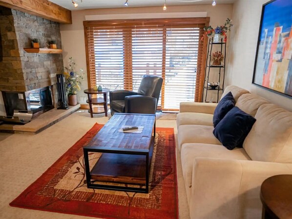 A living room with a fireplace, a sofa, a coffee table, and a large window with blinds.
