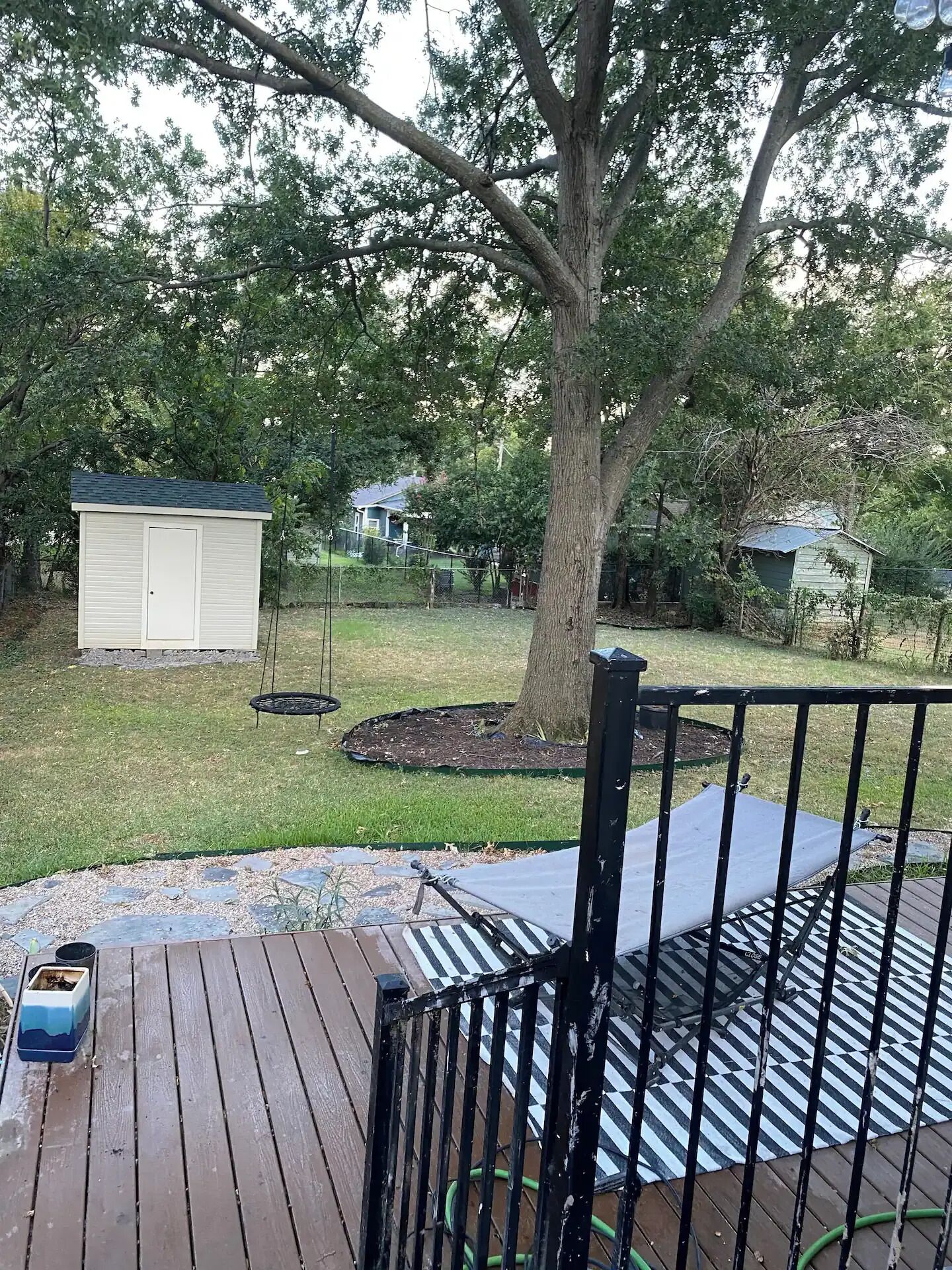 A wooden deck with a metal railing, a swing set, and a small shed in a backyard with a tree.