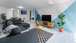 Een moderne woonkamer met een hoekbank, een televisie en een salontafel. Er staan potplanten en een kamerbreed tapijt.