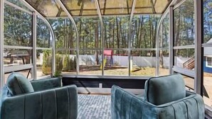 A conservatory with a glass ceiling, two green upholstered chairs, and a view of a forested area. - Strawberry Pines Retreat (Strawberry)