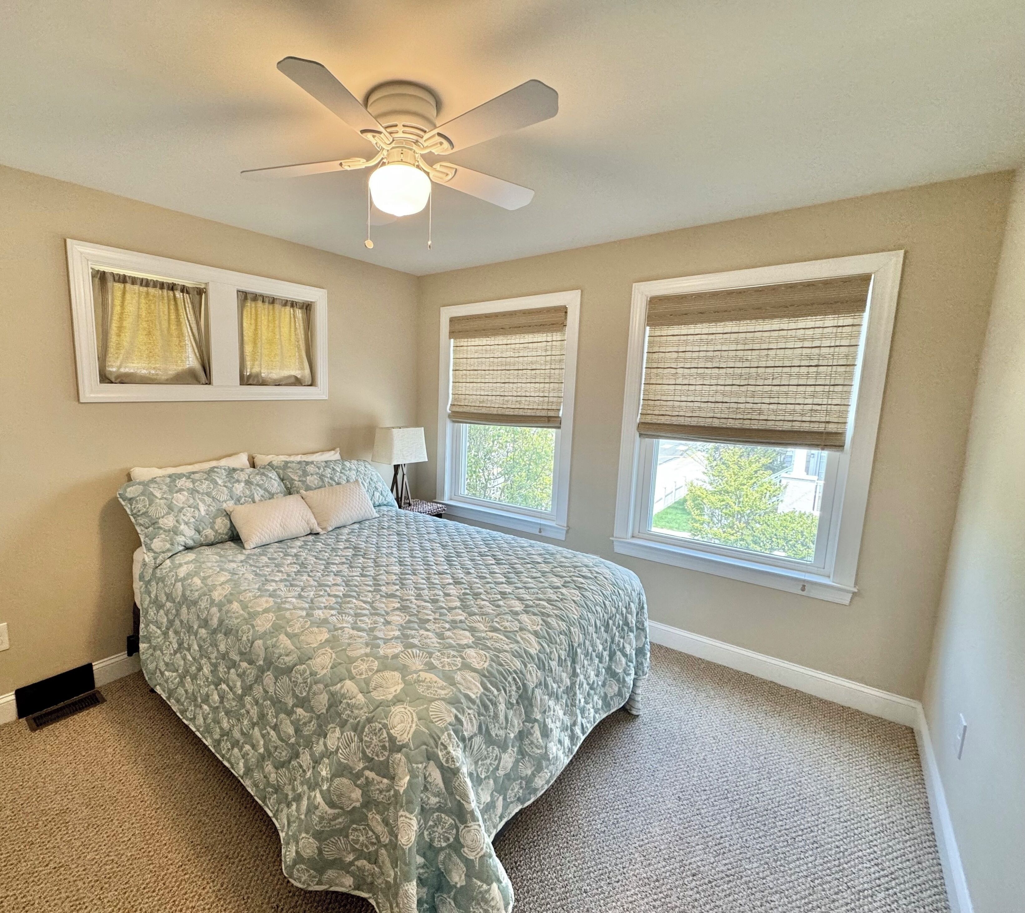 A bedroom with a bed, a ceiling fan, two windows with blinds, and a nightstand with a lamp.