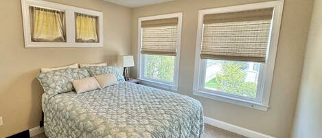 A bedroom with a bed, a ceiling fan, two windows with blinds, and a nightstand with a lamp.