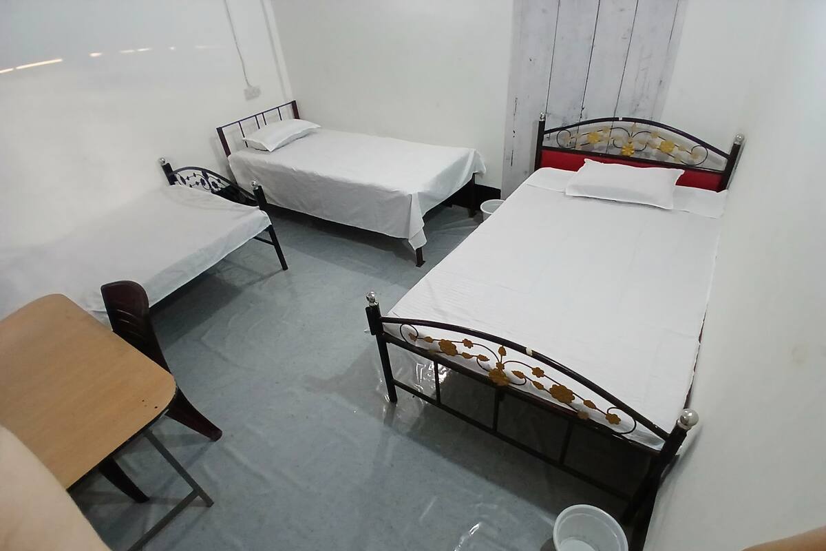 A dormitory room with three single beds, a wooden desk, and a chair.