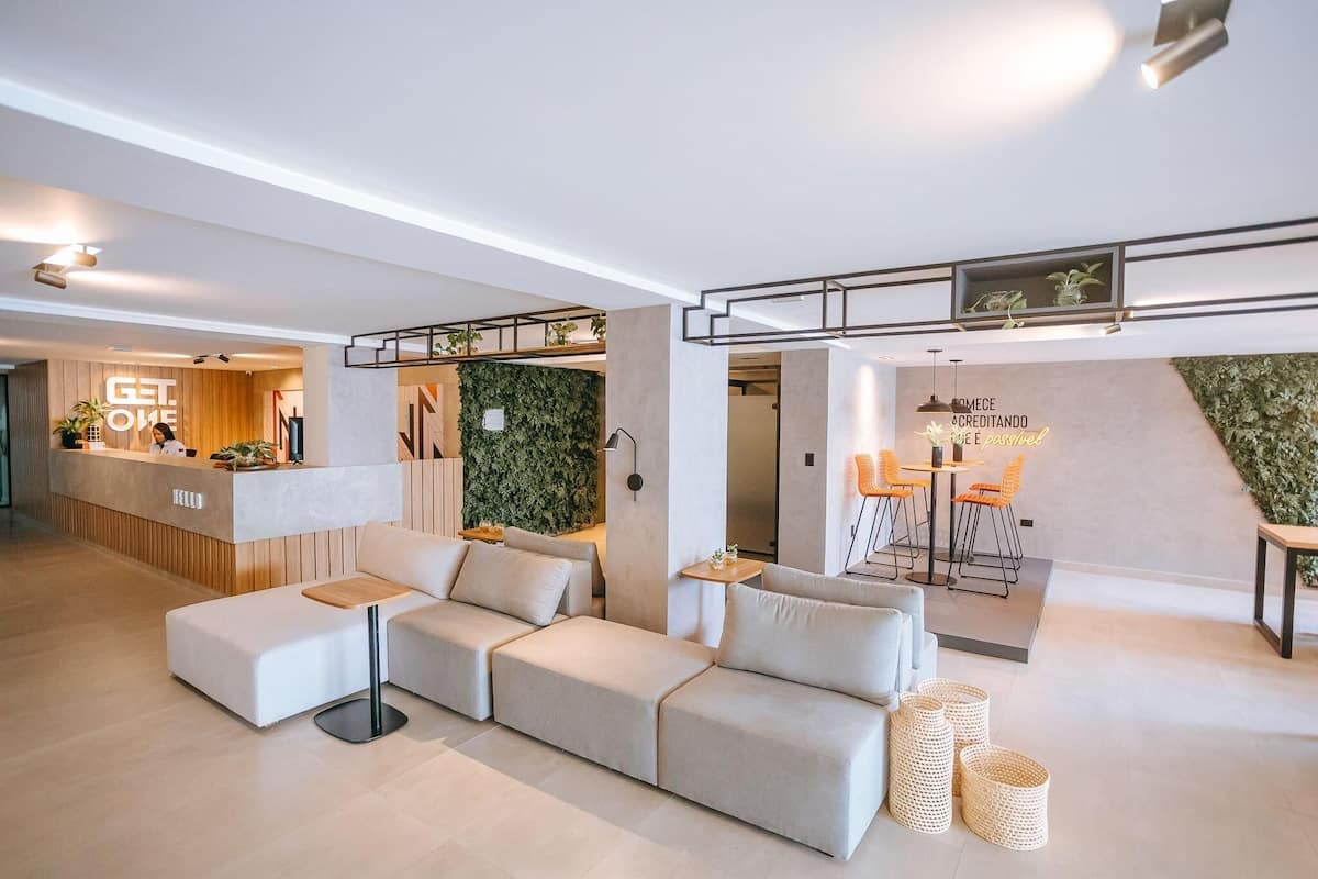 A modern interior with a reception desk, a green wall, a seating area with a sofa and ottoman, and a dining setup with chairs and a table.