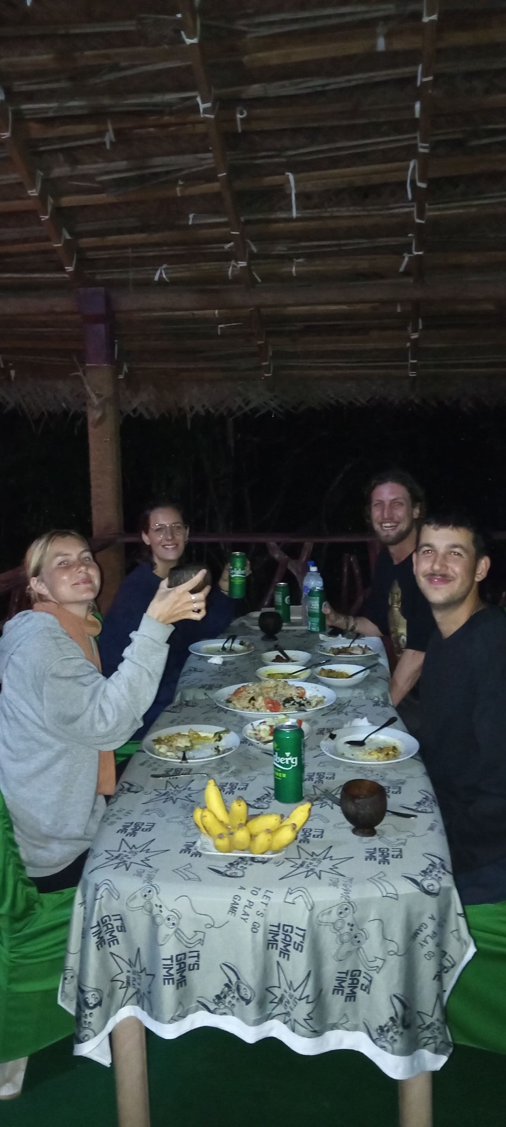 Eine Gruppe von Personen sitzt an einem Tisch mit einer Tischdecke, die Zeichentrickfiguren und den Schriftzug "It's game time" zeigt. Auf dem Tisch befinden sich Teller mit Essen, eine Schüssel, eine Banane und eine Dose Limonade.