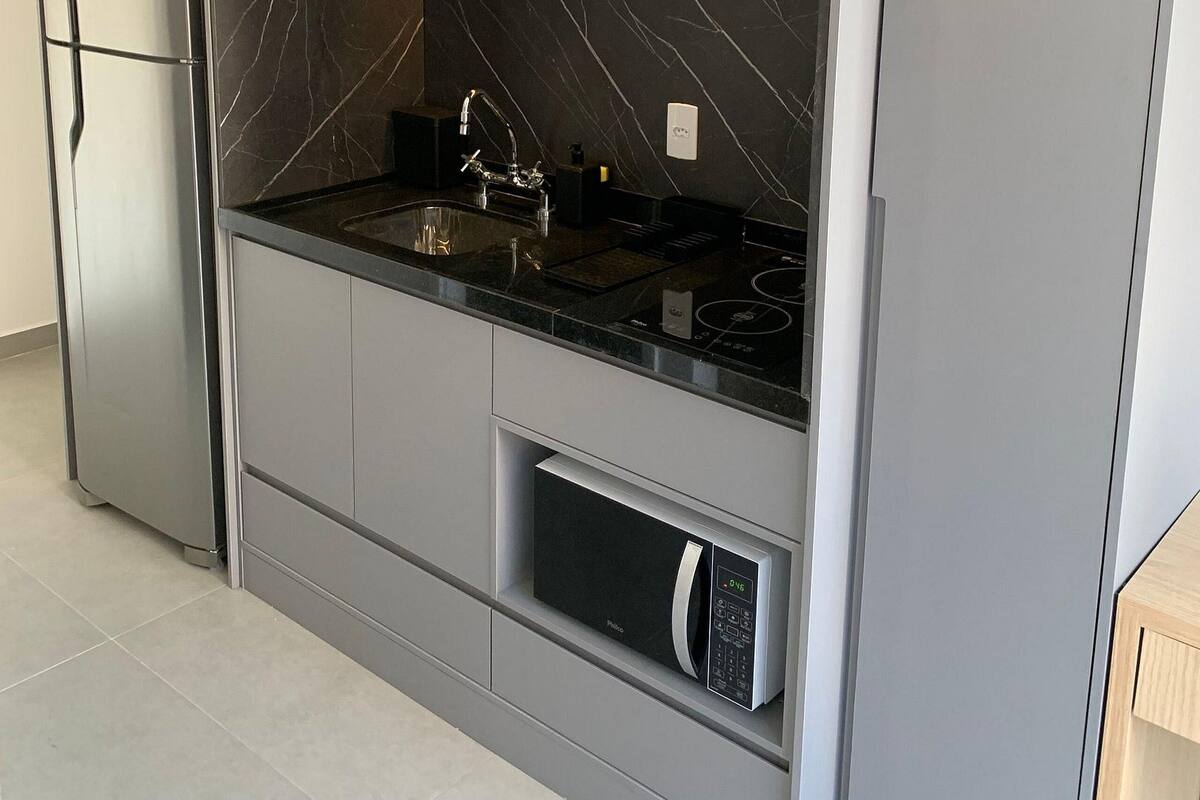 A modern kitchen with grey cabinetry, a black marble backsplash, a stainless steel refrigerator, a built-in microwave, and a black countertop with a sink and faucet.