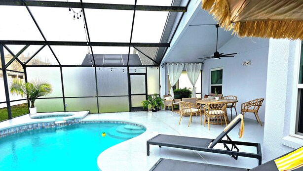 A pool area with lounge chairs and a table, surrounded by a glass enclosure with a ceiling fan and curtains.