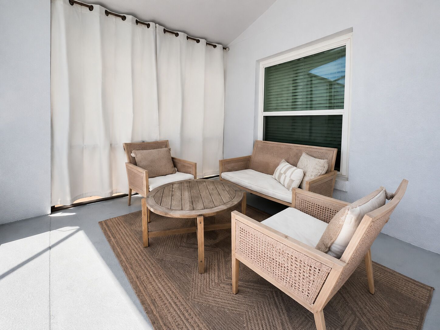 A sunlit outdoor seating area with wicker furniture, a round table, and a view of a building through a window.