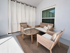 A sunlit outdoor seating area with wicker furniture, a round table, and a view of a building through a window.