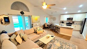 A spacious living area with a large sofa, a coffee table, and a dining table. There is a kitchen area with modern appliances and a large clock on the wall.