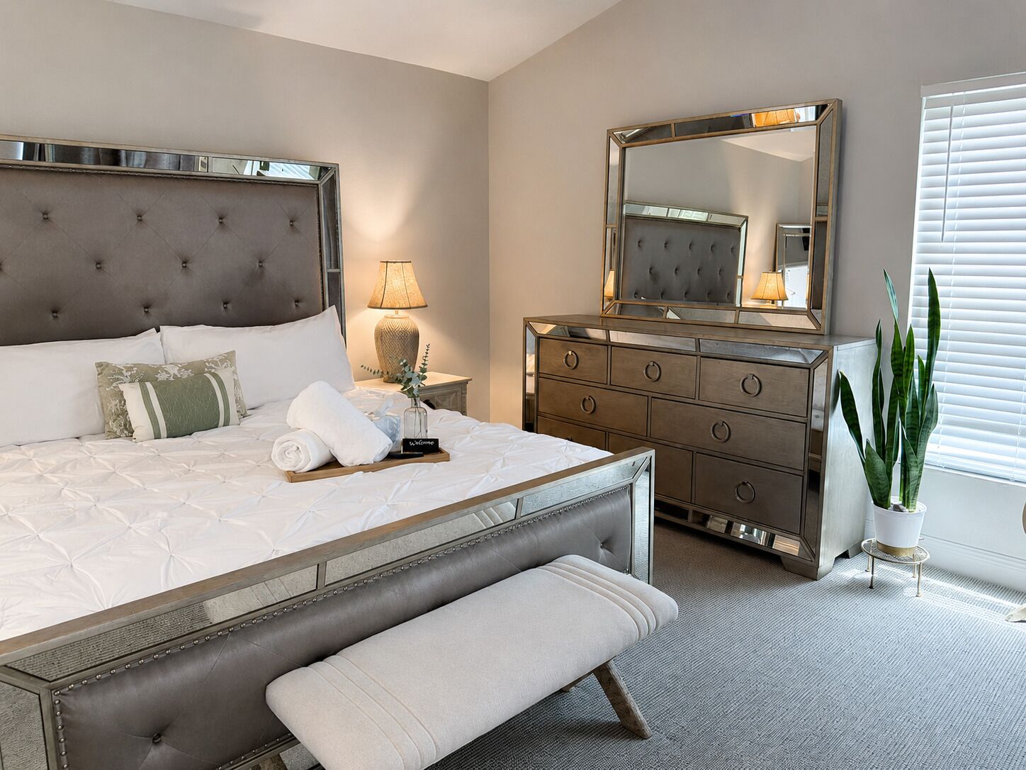 A bedroom with a large bed, a bedside table with a lamp, a dresser with a mirror, and a potted plant.