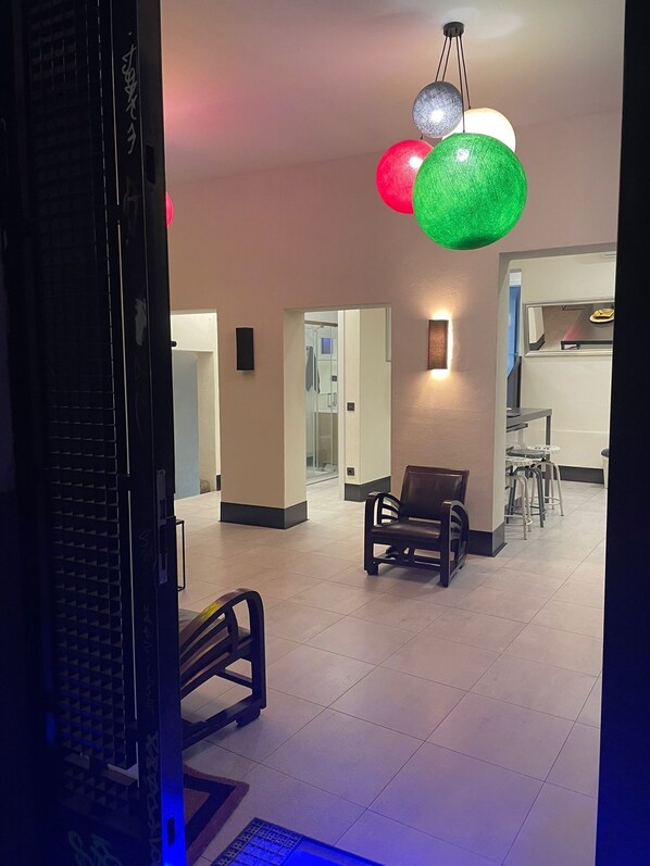 A hotel lobby with a reception desk, a waiting area with chairs, and a dining area with stools. There are decorative spherical lights hanging from the ceiling.