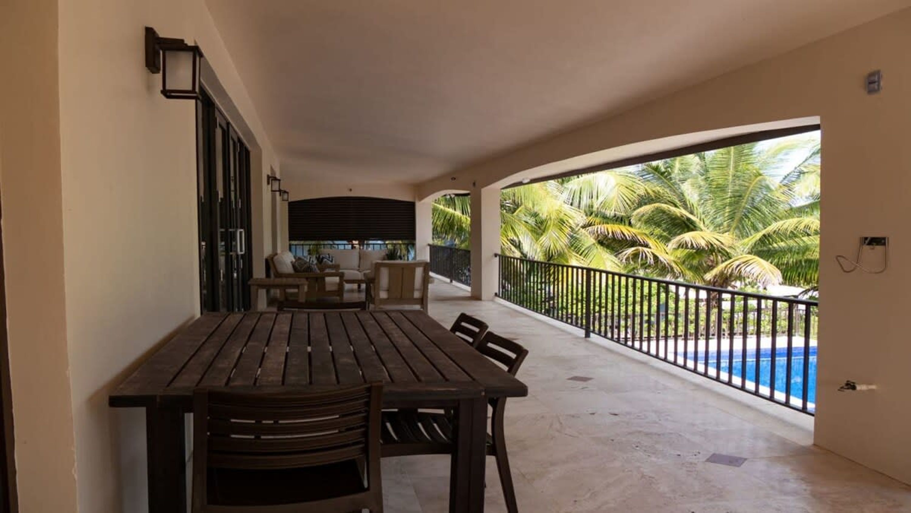 A covered outdoor area with a wooden table and chairs, a sofa, and a view of a swimming pool and palm trees.