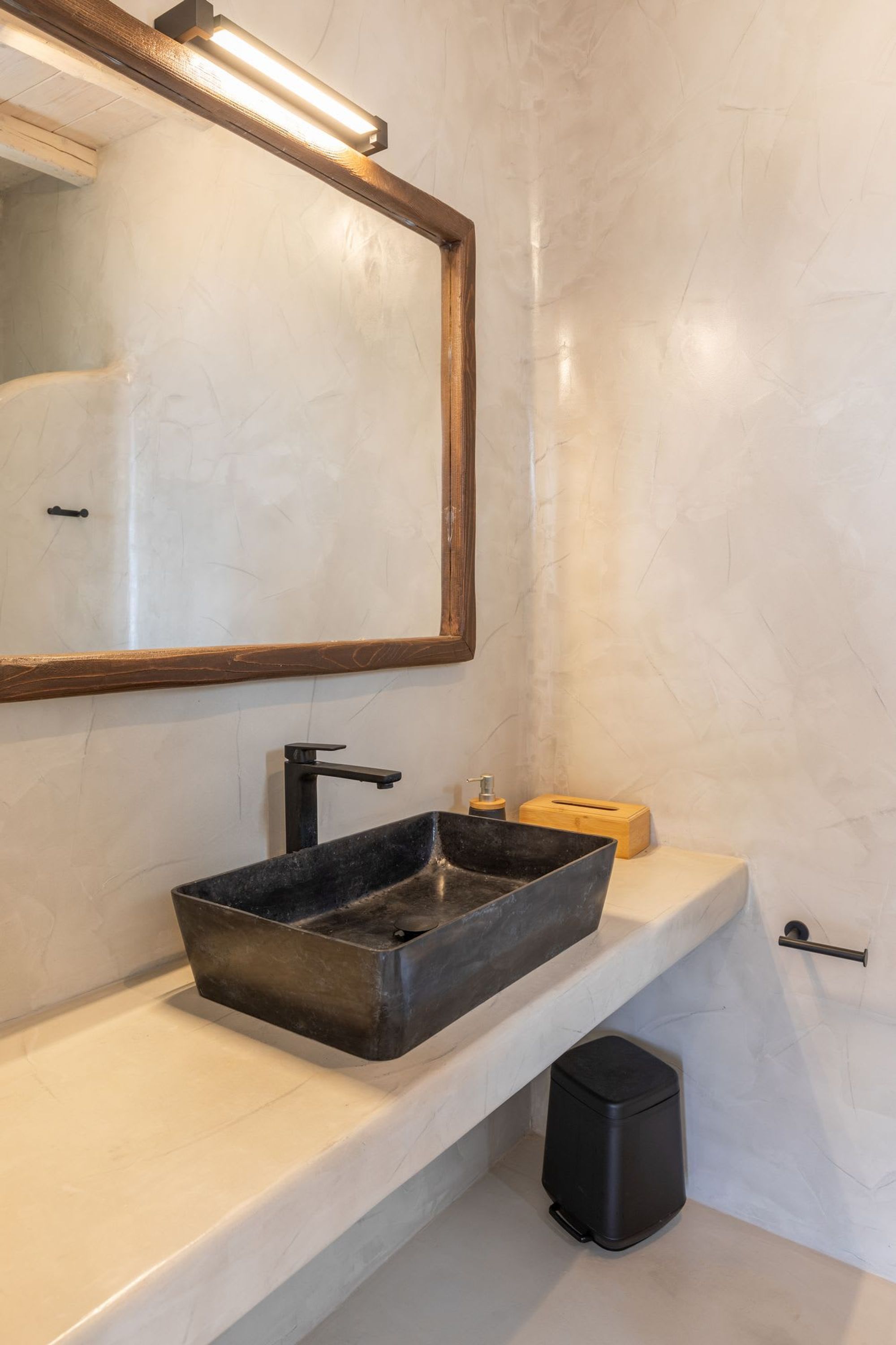 A bathroom with a large mirror, a black vessel sink, a wooden soap dish, and a black trash bin.