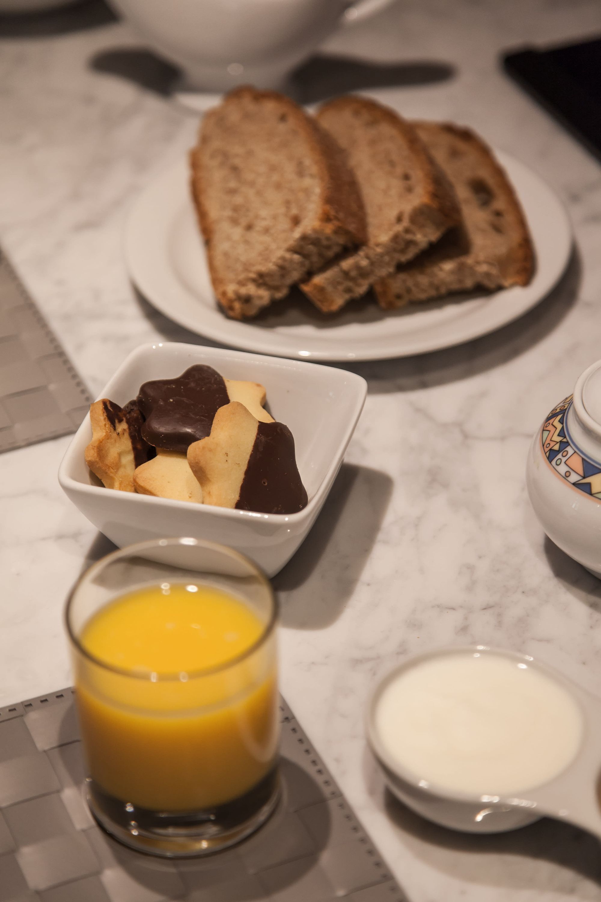 Une table dressée pour le petit-déjeuner avec des toasts, des biscuits enrobés de chocolat, un verre de jus d’orange et un petit bol de lait, le tout posé sur un comptoir en marbre.