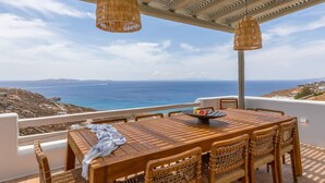 A wooden table with wicker chairs and a wicker pendant light, overlooking a coastal landscape. - Modern Mykonos Villa Private Pool Stunning View (Mykonos)