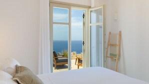 A bedroom with a large bed, a view of the sea through an open door, and a wooden chair outside. - Modern Mykonos Villa Private Pool Stunning View (Mykonos)