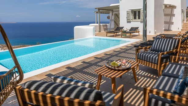 A poolside area with lounge chairs, a table with a fruit bowl, and a view of the sea. - Modern Mykonos Villa Private Pool Stunning View (Mykonos)