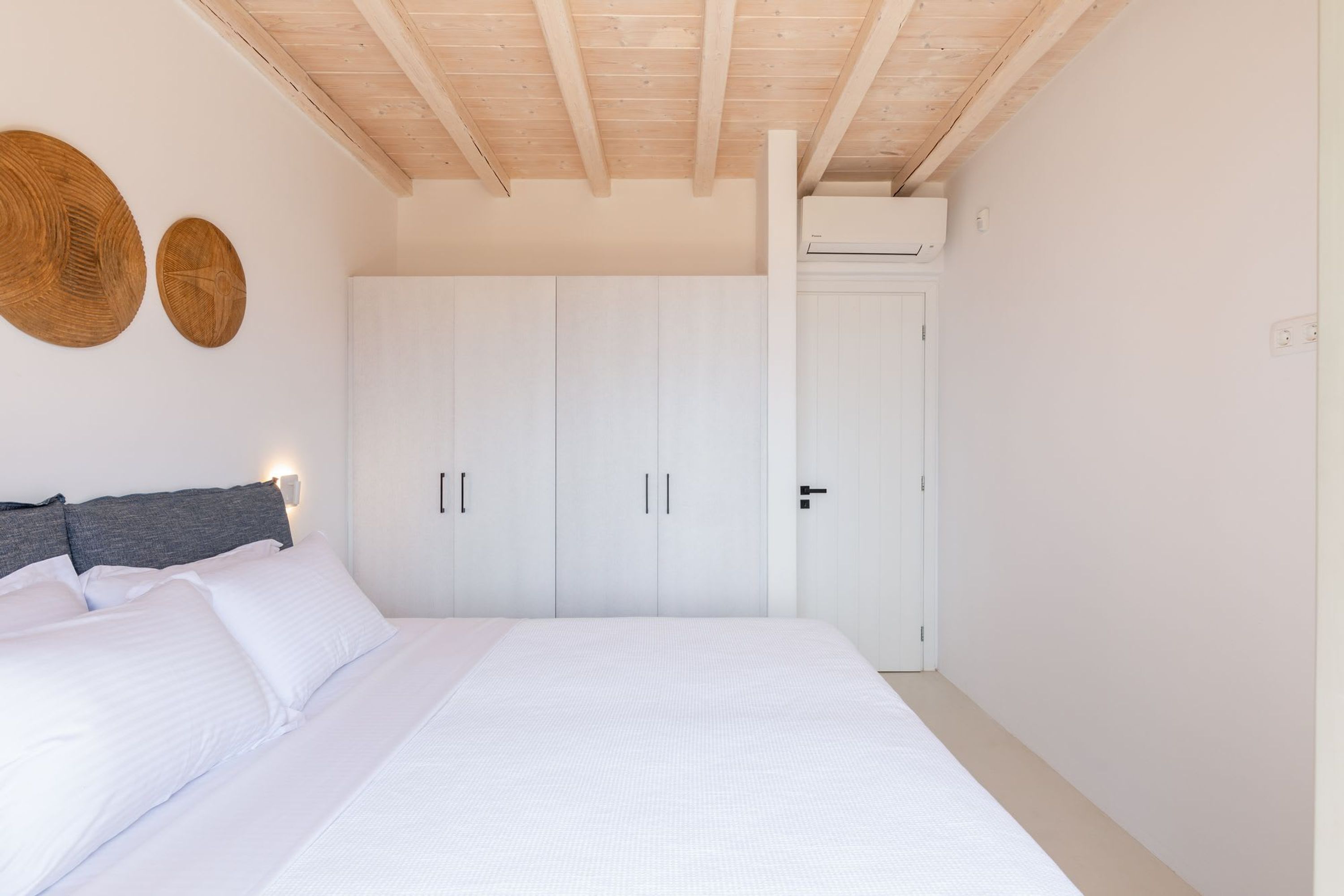 Une chambre avec un grand lit, des murs blancs, des poutres en bois au plafond et une armoire à plusieurs portes.