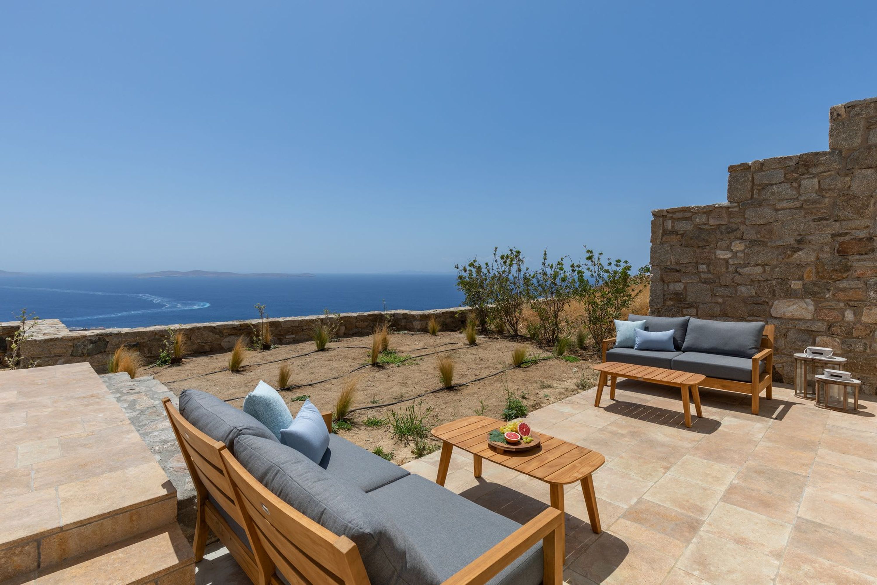 A patio with a stone wall, a wooden sofa, a wooden table, and a small round table with a tray of fruit.