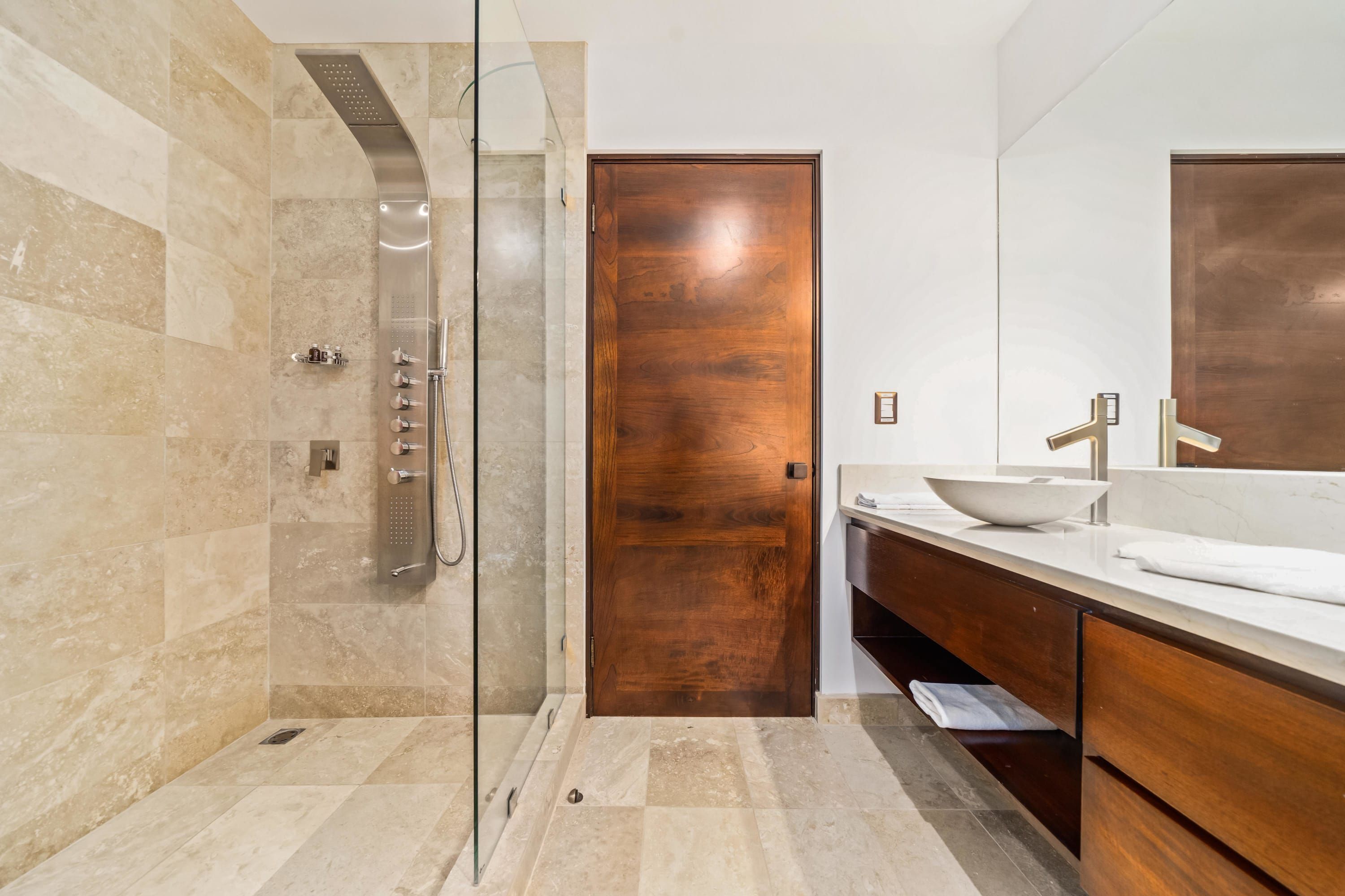 A modern bathroom with a glass-enclosed shower, a wooden door, and a vanity with a sink and mirror.