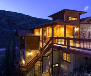 A multi-story log cabin with a wooden deck and staircase, surrounded by a mountainous landscape.